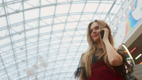 mujer joven sonriente con camiseta azul de pie en el centro comercial usando un teléfono inteligente, navegando, leyendo noticias, hablando con amigos