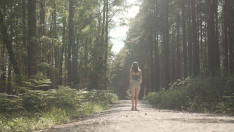 young woman walking down forest road on summer day, following rear shot