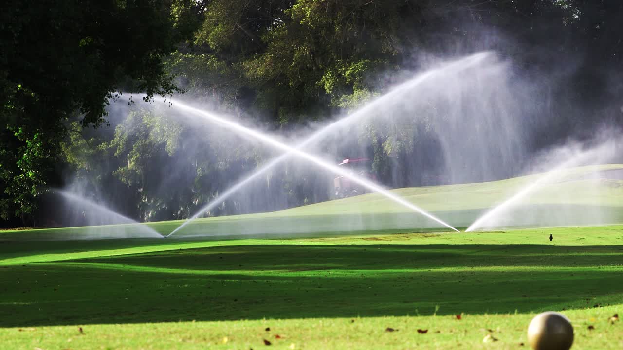 Riding lawn mower in background of golf course as sprinklers water the green grass