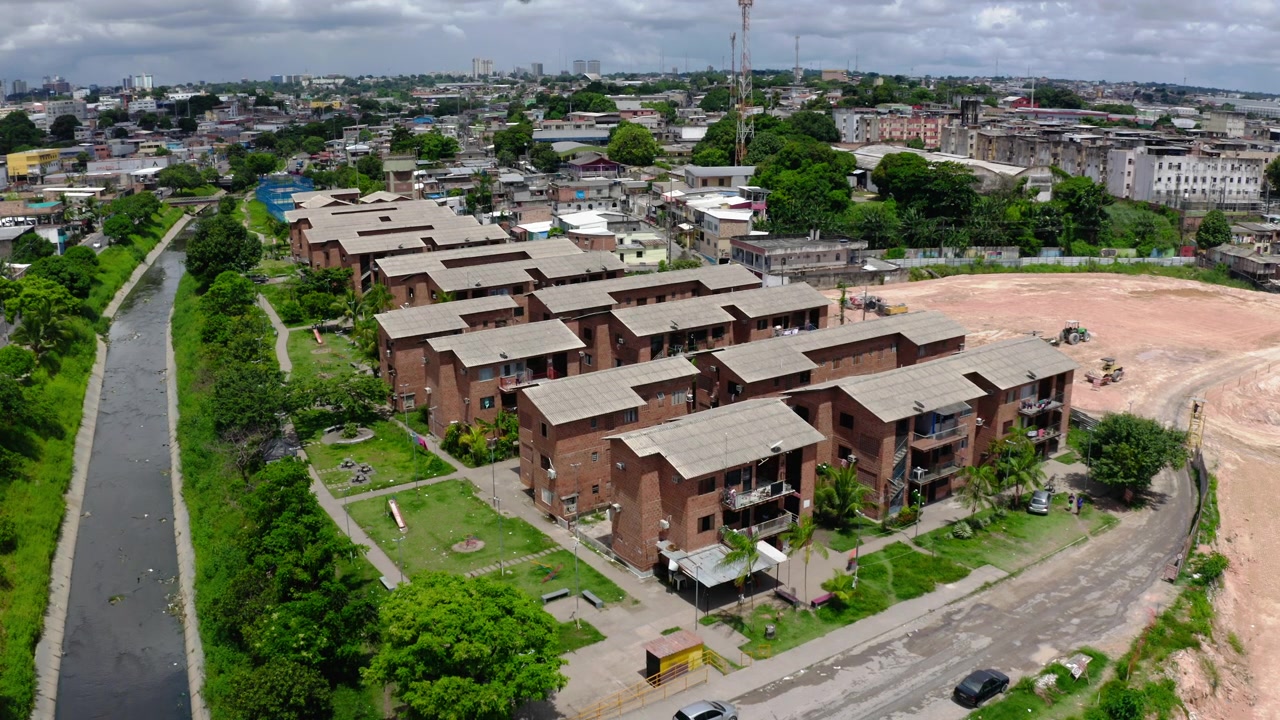 Static aerial drone view of rows of brick social housing blocks of a conjunto habitacional beside the canalised igarape in Manaus, lawns, palms, parked cars, graded lot, city skyline, sunny, Brazil.