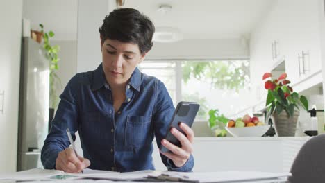 Caucasian-woman-taking-notes-and-calculating-finances-using-smartphone-at-home