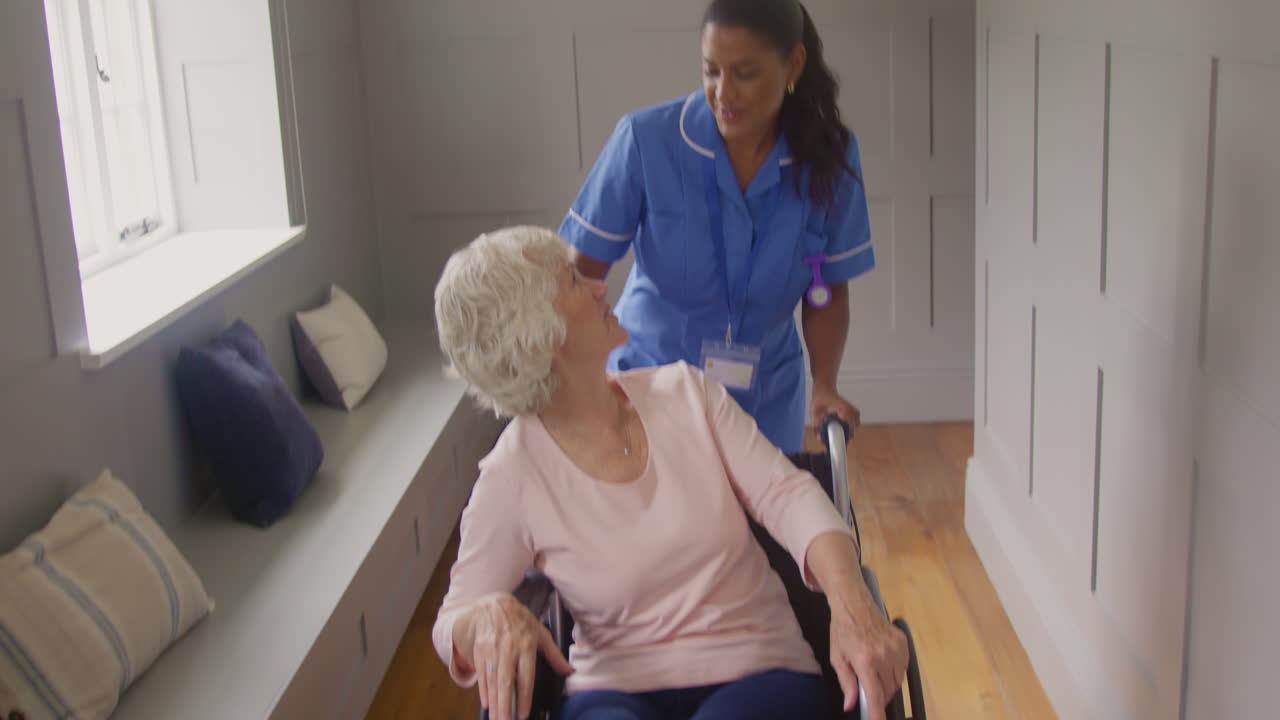 mujer mayor en casa siendo empujada en silla de ruedas por una trabajadora de atención femenina en uniforme