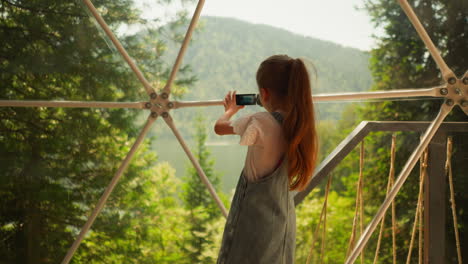 niño toma video del lago y la montaña en glamping. niña pequeña sostiene la cámara de vídeo por la ventana panorámica en el hotel ecológico. niño juega cámara en el complejo