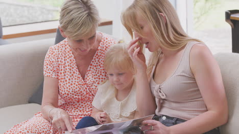 Granddaughter-Sitting-On-Sofa-With-Mother-And-Grandmother-At-home-looking-At-Photo-Album-Together