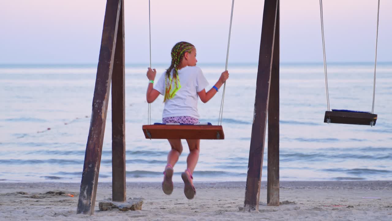 Girl enjoying a beautiful sea landscape on a swing. Young female resting alone near water. Back view of a girl swinging against the sea background in the evening.