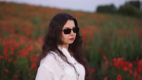 beautiful-dark-haired-girl-in-a-field-of-wildflowers-and-red-poppies,-wearing-a-dress-and-smiling-while-putting-on-sunglasses