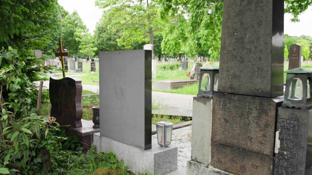 Lapidi al Zentralfriedhof, Vienna, Austria, mentre la telecamera panoramica verso destra dietro monumenti con alberi verdi, prato e sentiero. Atmosfera tranquilla e pacifica del cimitero in estate.