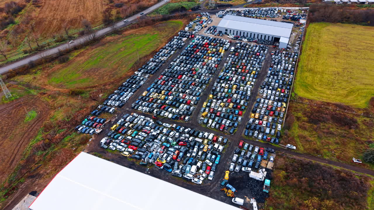 Vista aérea de gran altura de un gran desguace de automóviles. Toma amplia de un depósito de chatarra de coches con filas de vehículos junto a un almacén industrial y una carretera rural