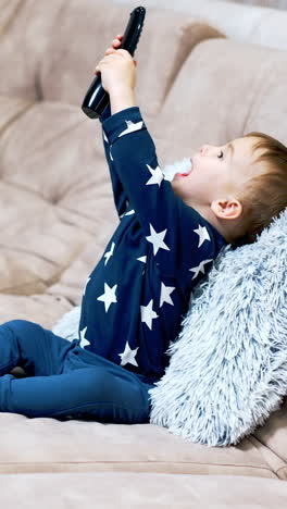 beautiful little toddler in blue clothes sitting on a big sofa. lovely kid is playing with a remote control. side view. vertical video