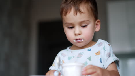 lovely cute male kid with dairy on his lips. baby boy eating yogurt from cup. low angle view. blurred backdrop.