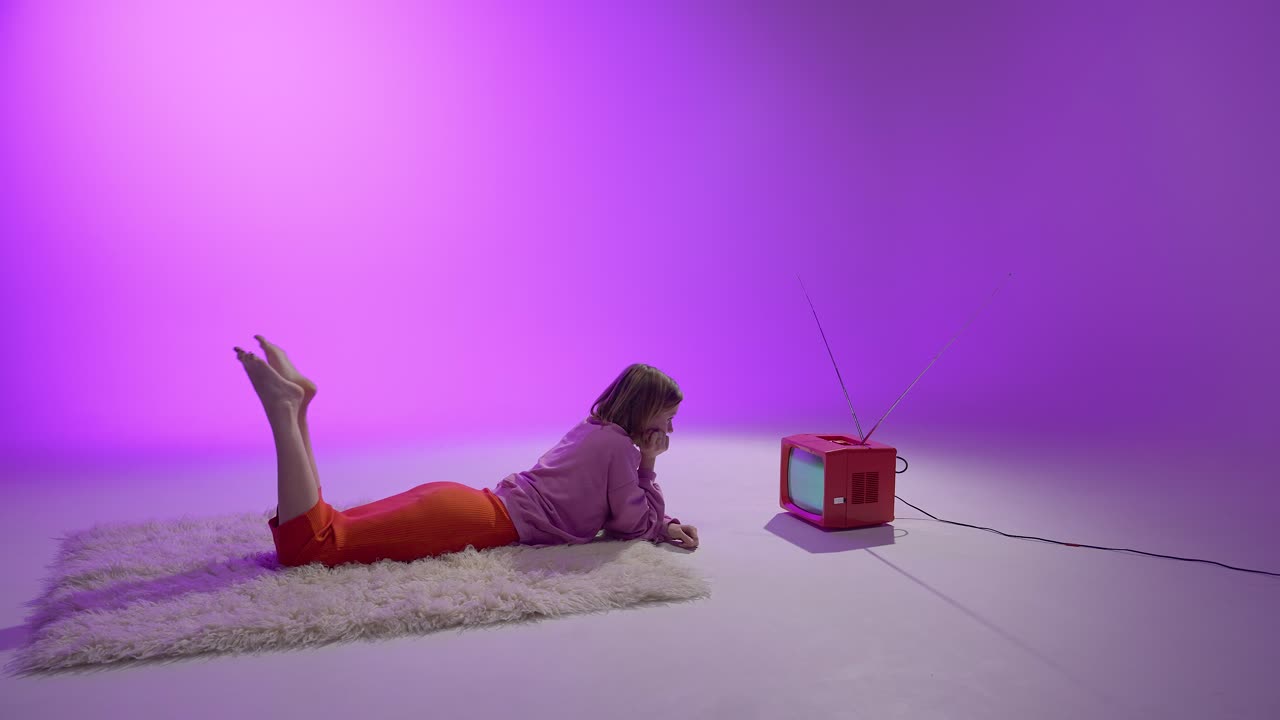 A young woman lies comfortably on a soft rug in a purple-lit studio, reaching to adjust a red retro TV with static. The scene blends nostalgia with modern color aesthetics for a vintage-inspired mood