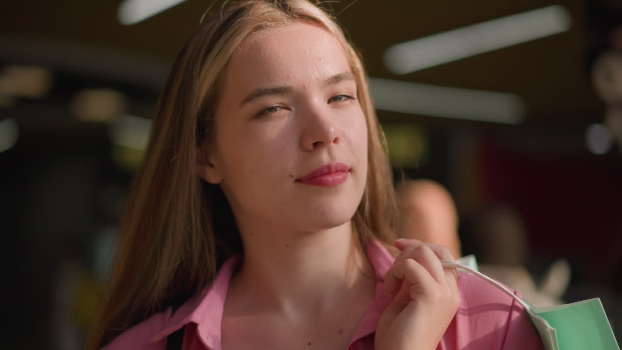 매력적인 미소를 짓는 우아한 젊은 여성은 그녀의 뒤에 가방을 들고 카메라를 자신감 있게 쳐다보고, 배경은 불분명한 물체로 흐릿해지고 부드럽고 반이는 효과를 제공합니다.