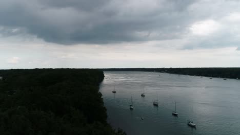 Ariel-drone-footage-of-several-sailboats-sailing-in-a-line,-with-dark-trees-on-the-coastline