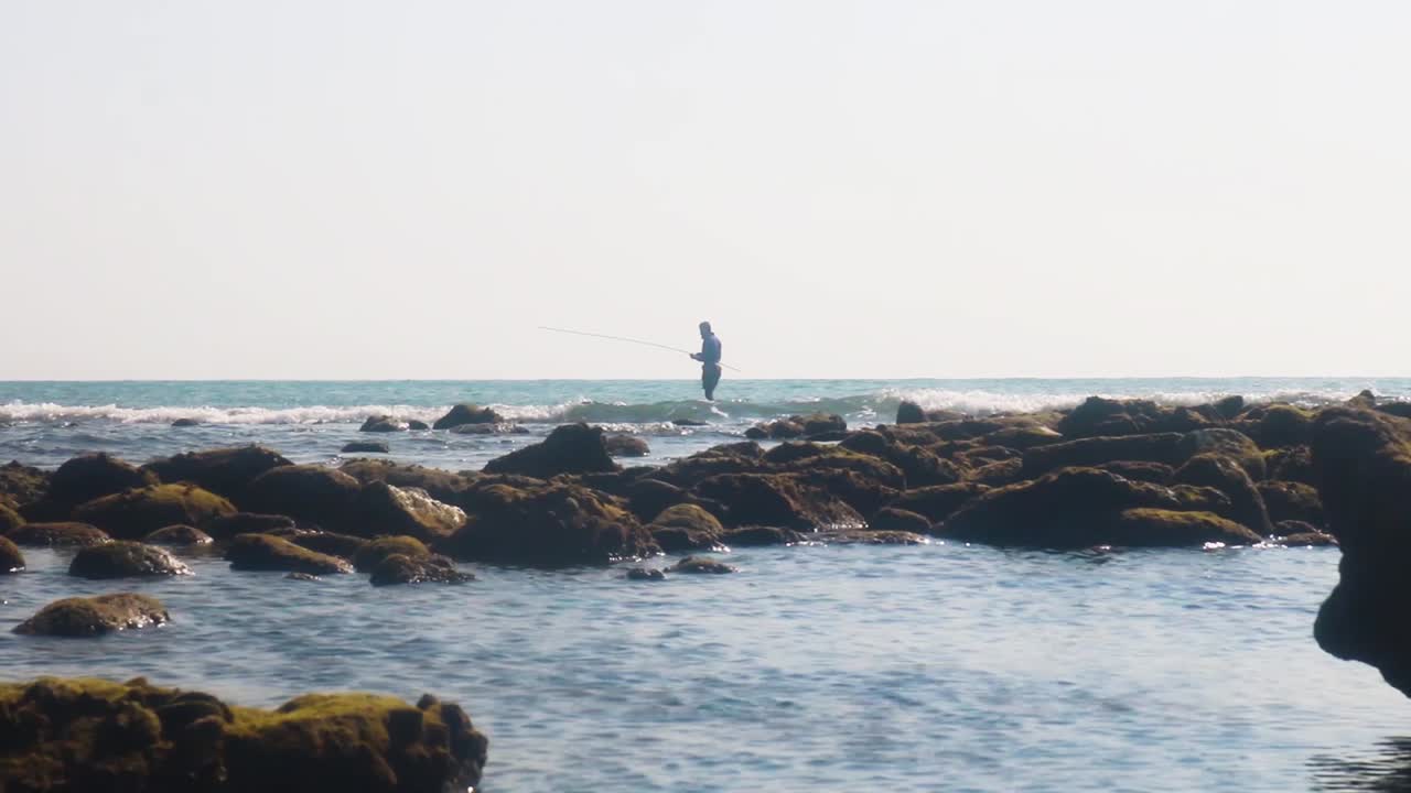 Pescatore solitario che pesca su una costa tropicale rocciosa, piccole onde oceaniche
