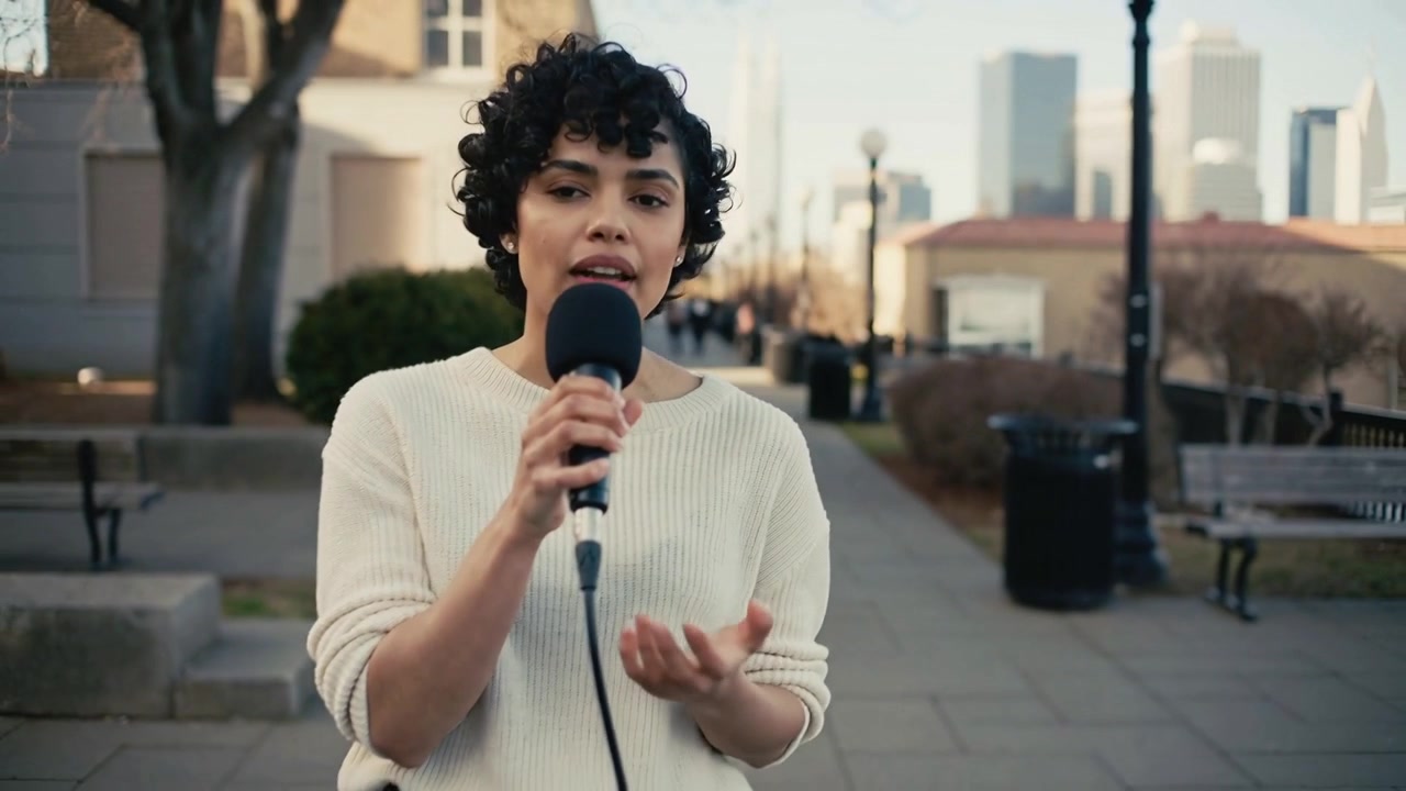 Mujer hispana cantando con suéter claro y pantalones oscuros, sosteniendo un micrófono con cable mientras la cámara panea la plaza.
