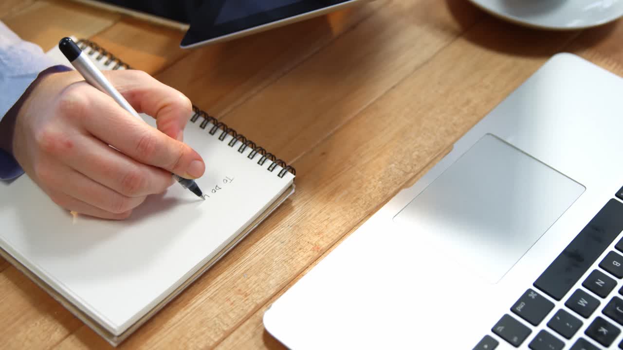 Hand of businesswoman writing on notebook at desk with digital tablet and laptop on table