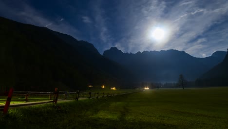 Timelapse-of-Alpine-valley-by-night-under-full-moon,-illuminated-farmhouse,-cottage,-full-moon,-clouds-and-stars-in-sky,-light-trails-of-cars-passing-on-road