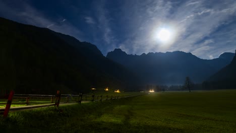 timelapse of alpine valley by night under full moon, illuminated farmhouse, cottage, full moon, clouds and stars in sky, light trails of cars passing on road