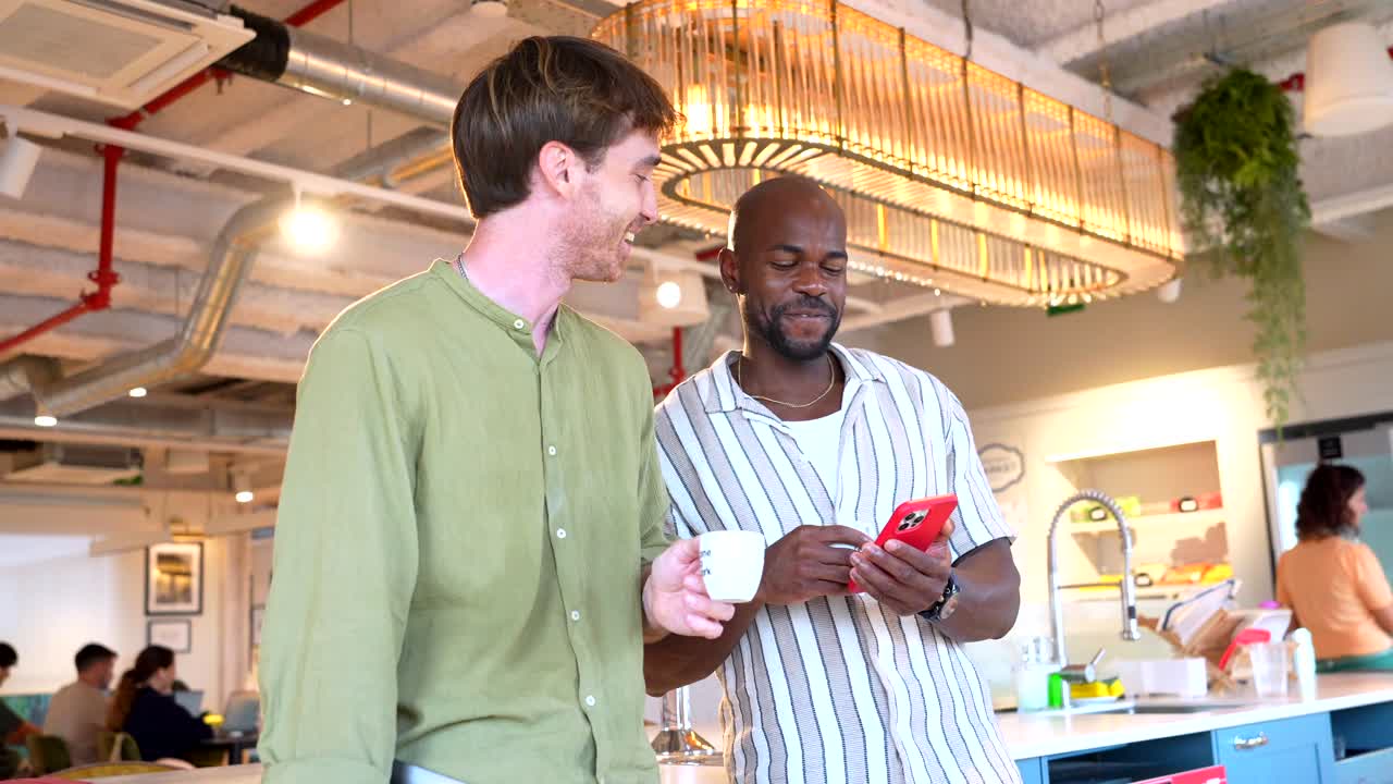 Two Diverse Colleagues Chatting and Looking at a Smartphone in Office Breakroom