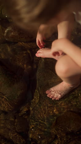 little girl washes feet with river water sitting on rock. weekends in wilderness by clear mountain river on sunny day. concept of relaxation without gadgets