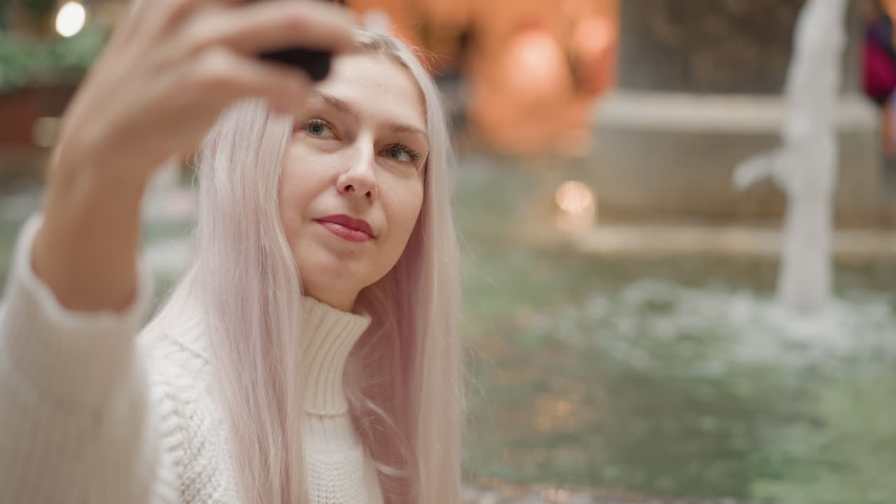 portrait of urban woman sits by flowing fountain taking selfie, candid expression visible against blurred mall interior lighting, capturing tranquil leisure moment in vibrant space