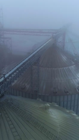 dusty tops of grain elevators at modern agricultural plant. flying above powerful silo complex on foggy winter day. aerial view. vertical video
