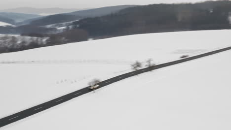 Tracking-Aerial-Shot-Of-Car-Driving-On-Road-in-Pristine-Winter-Scene,-Snow-Covered-Landscape-In-Europe