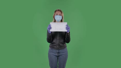 retrato de una hermosa mujer joven con máscara médica está mirando a la cámara y sosteniendo un cartel con las palabras quedarse en casa. rubia con cabello suelto en una chaqueta negra y vaqueros en una pantalla verde en el estudio. protección de la salud concepto de virus corona