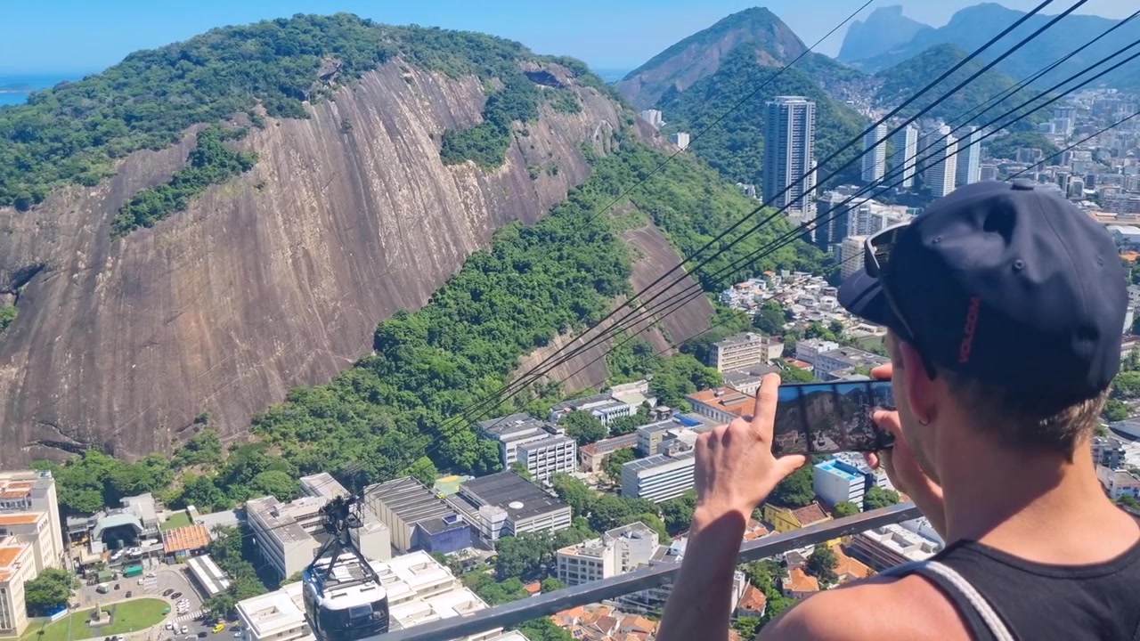 Turista maschio fotografa l'iconica funivia del Pan di Zucchero a Rio de Janeiro, Brasile. Rigogliose colline tropicali e tempo soleggiato creano un'atmosfera vivace in questa popolare attrazione turistica