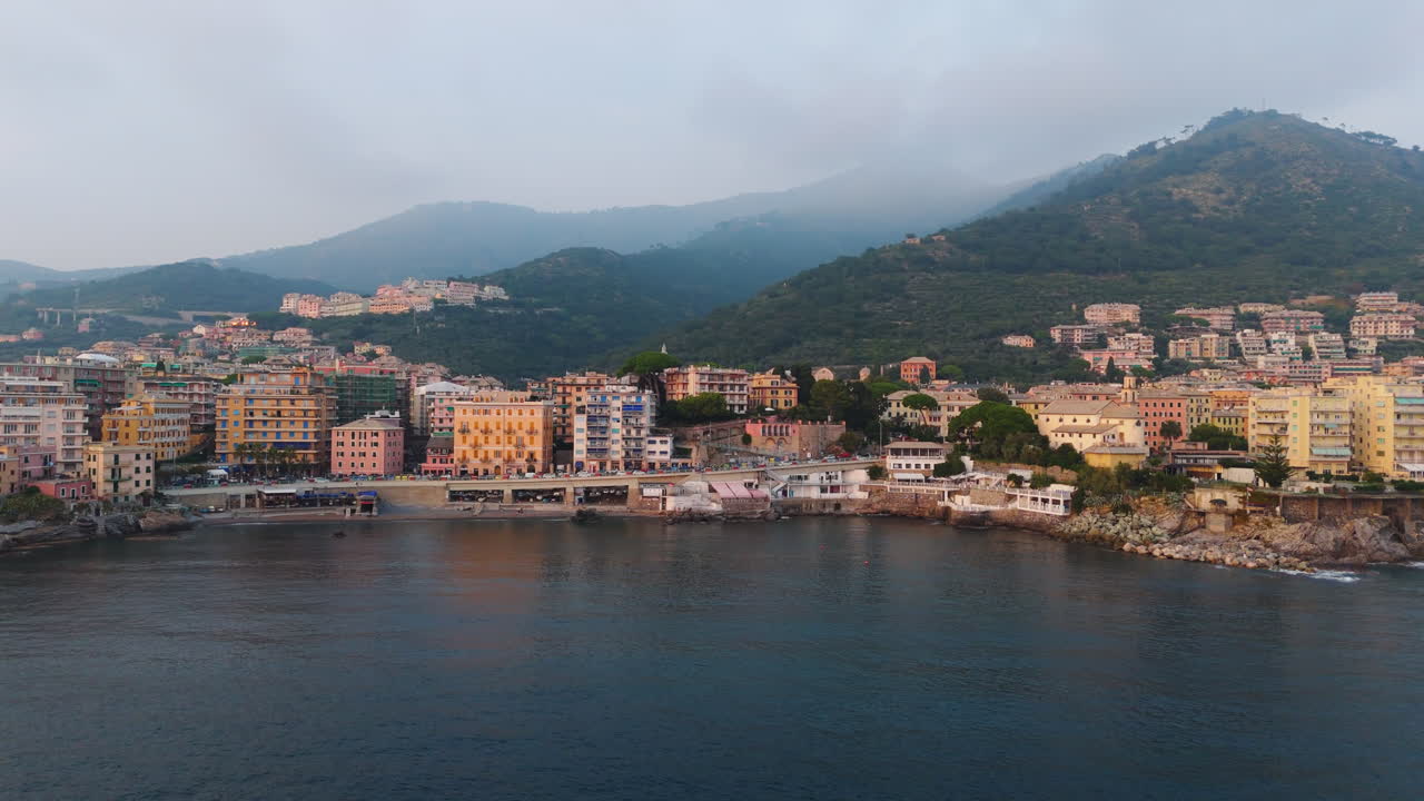 Il drone si allontana lentamente su una baia ligure, mostrando edifici colorati lungo la costa rocciosa, con montagne che si ergono sullo sfondo