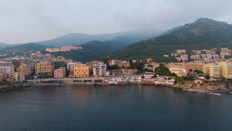 the drone slowly pulls back over a ligurian bay, showing colorful buildings along the rocky shore, with mountains rising in the background