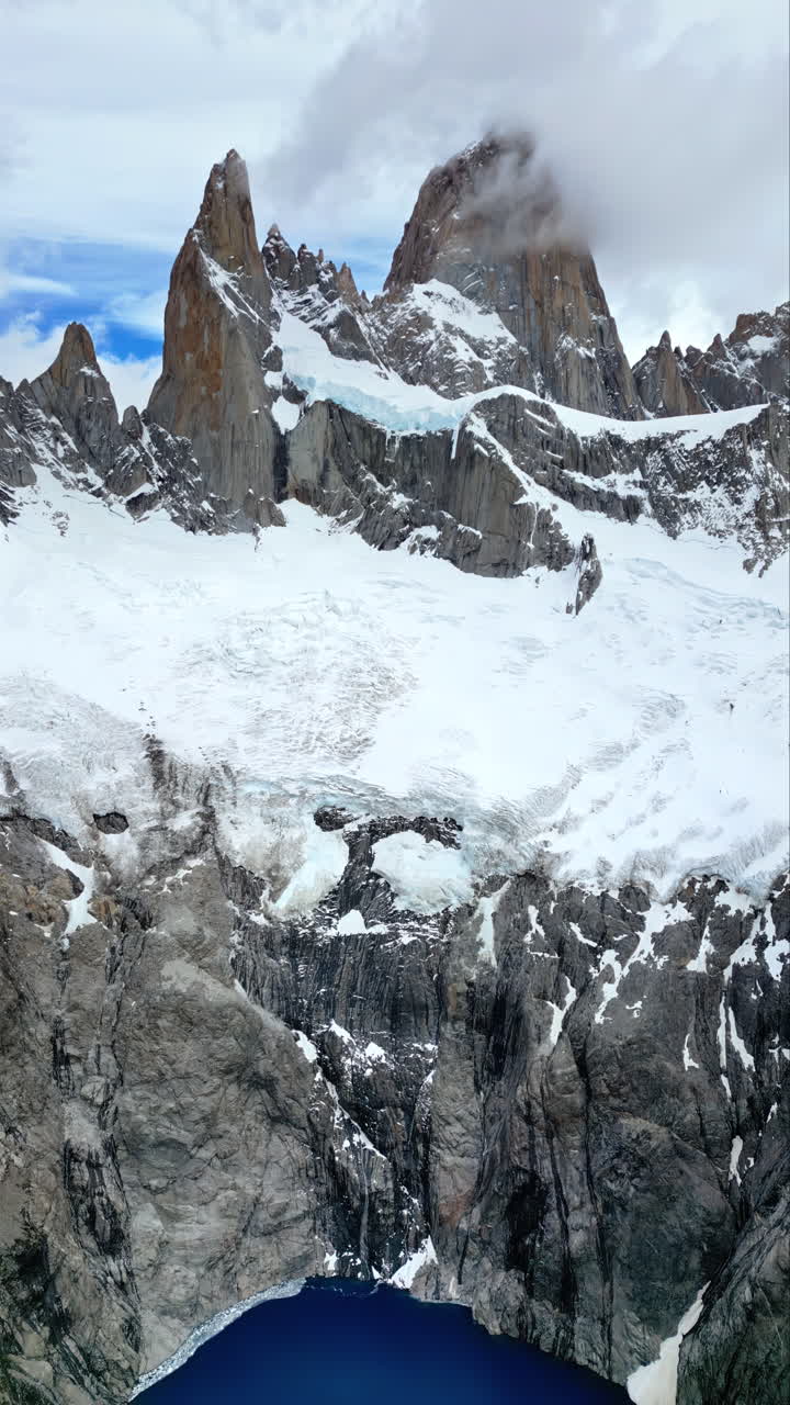 Vista aerea con drone delle maestose cime del Monte Fitz Roy che si ergono sopra un lago glaciale blu intenso, circondato da scogliere rocciose. Verticale