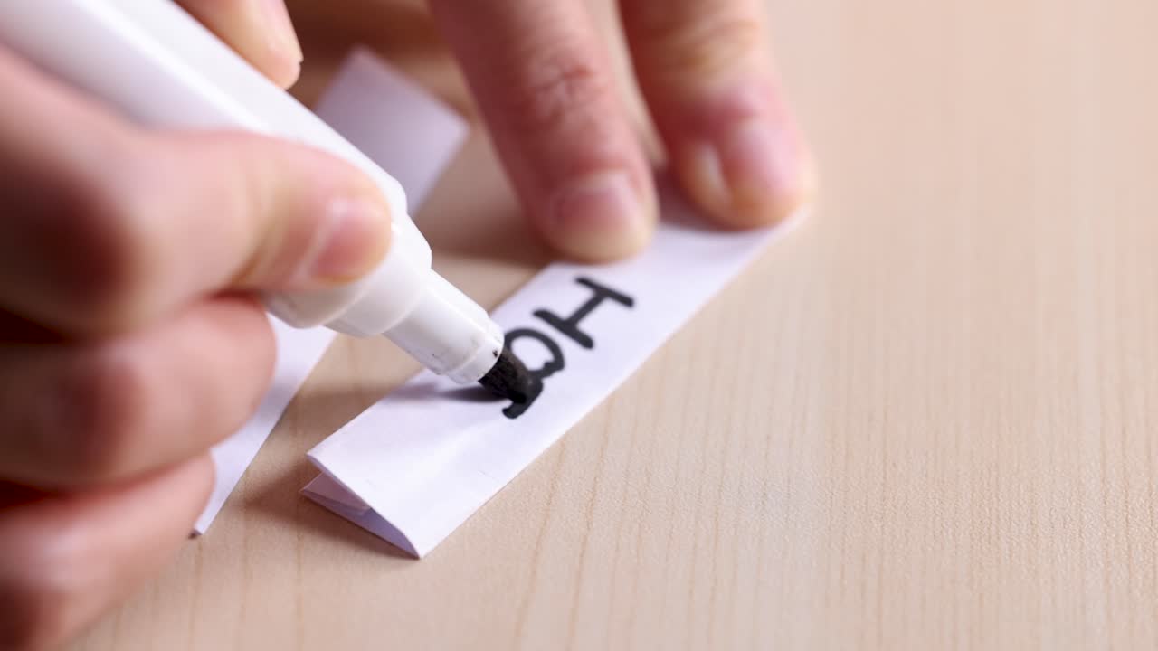 Close-up of hand writing 'Hot' with black marker on folded paper strip, natural lighting