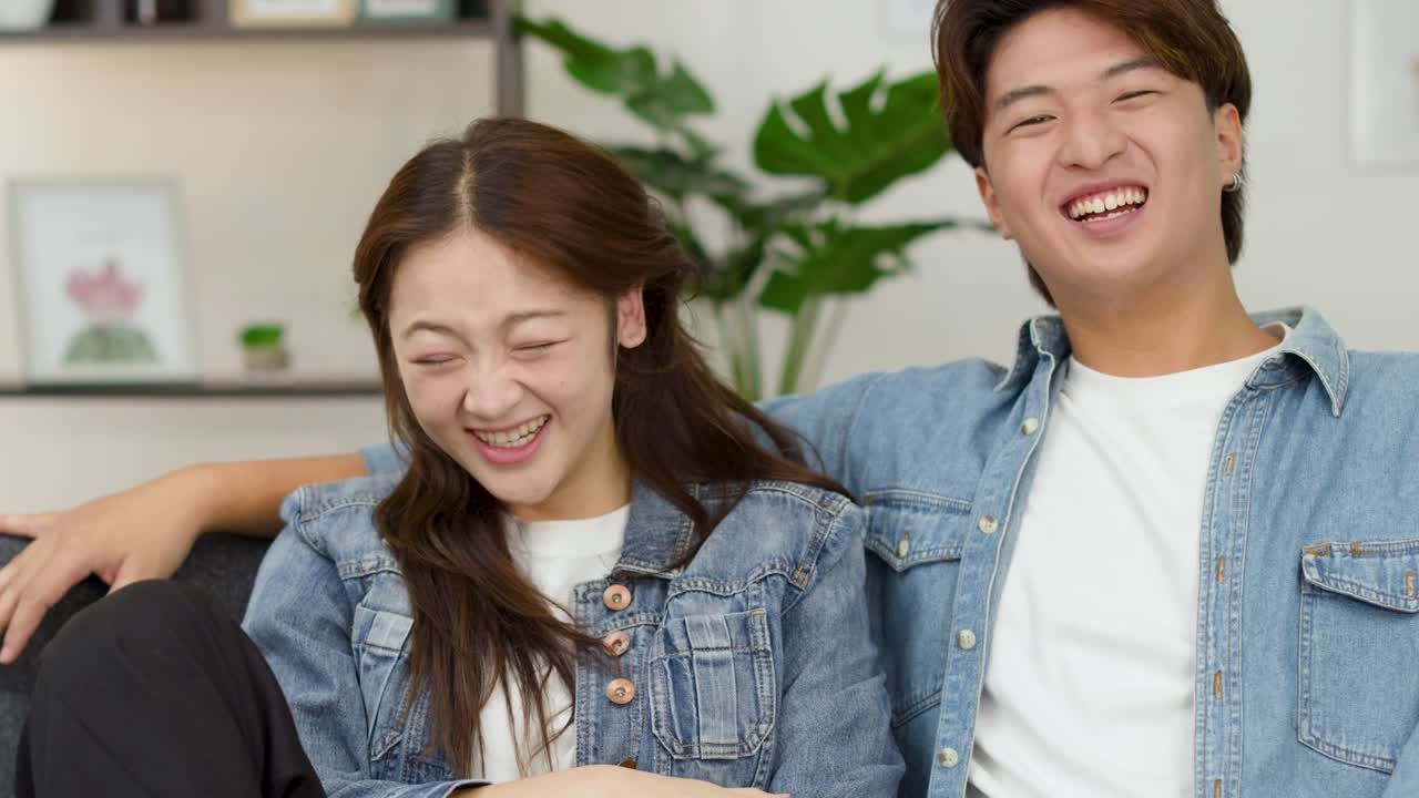 ソファで笑い合うデニムジャケット姿のアジア系の幸せそうなカップル、ミディアムショット、柔らかい室内照明。