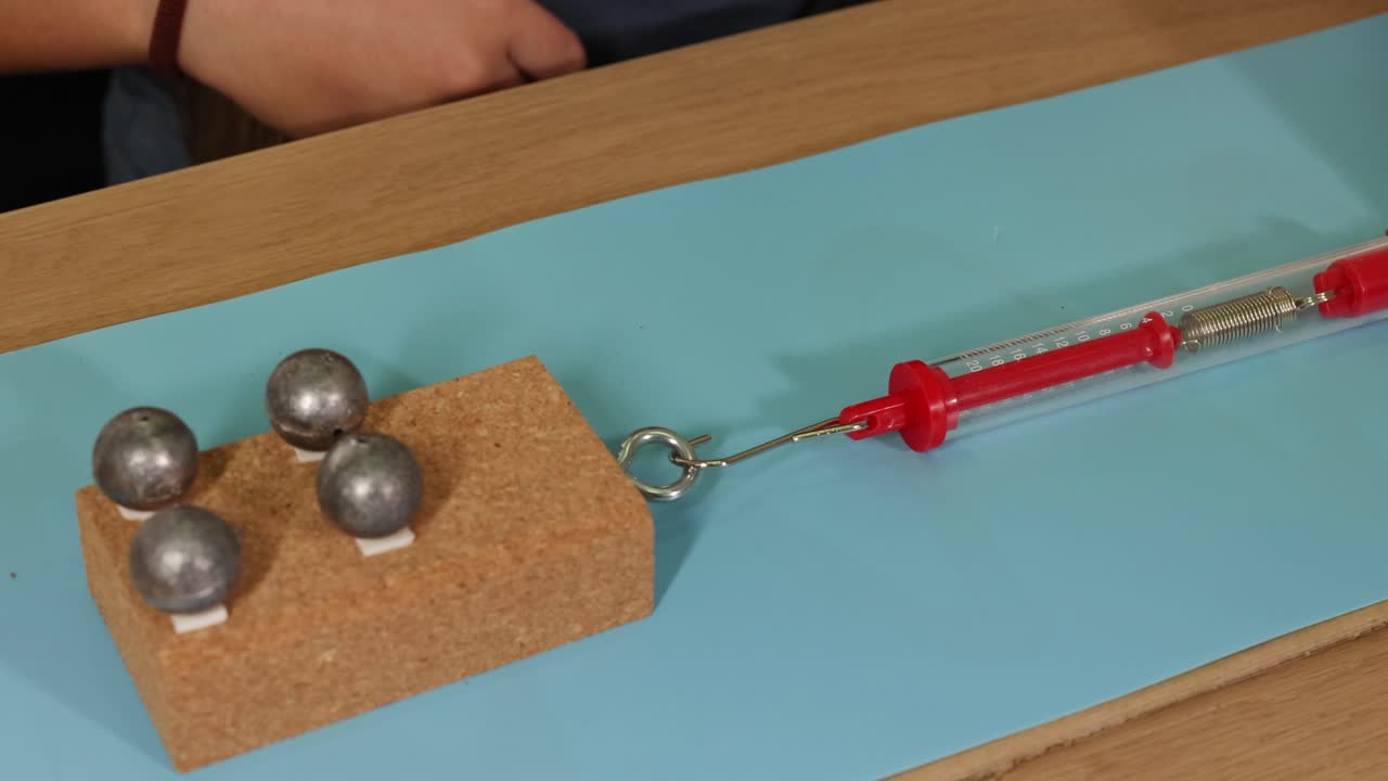 Hands manipulate weights on cardboard using a spring scale, illustrating friction and force dynamics on a smooth surface
