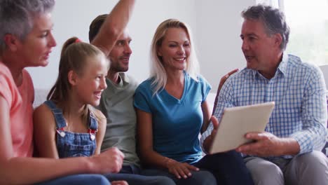 Smiling-caucasian-parents,-grandparents-and-granddaughter-sitting-on-couch-looking-at-tablet