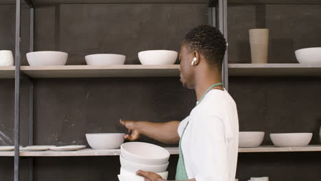 Male-Clerk-With-Wireless-Earbuds-Stocking-Ceramic-Bowl-On-The-Shelf-In-The-Pottery-Store