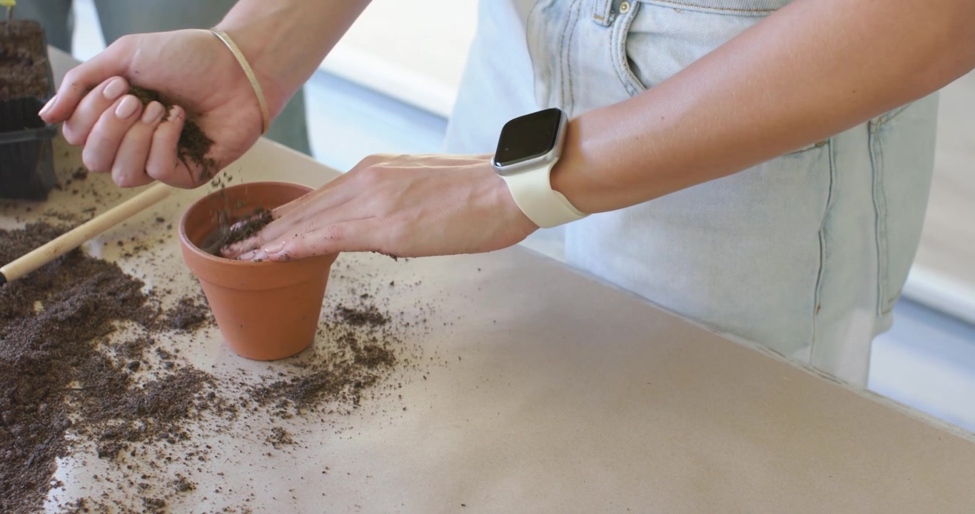 Donna che raccoglie e compatta il terriccio dal mucchio a sinistra sul tavolo, preparando un vaso di terracotta per la semina.