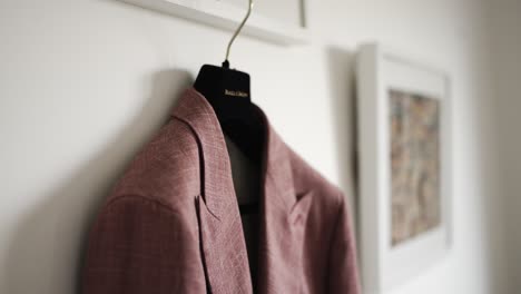 chaqueta de traje rosa colgada en un colgante negro contra una pared blanca, con arte enmarcado en el fondo