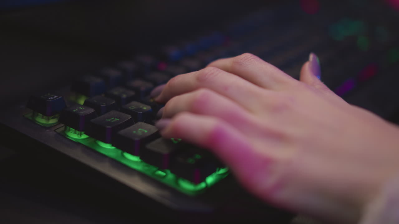 Hand view of woman with polished nails typing on rgb mechanical keyboard illuminated by colorful ambient light, showcasing modern gamer or streaming setup with close attention to tech detail