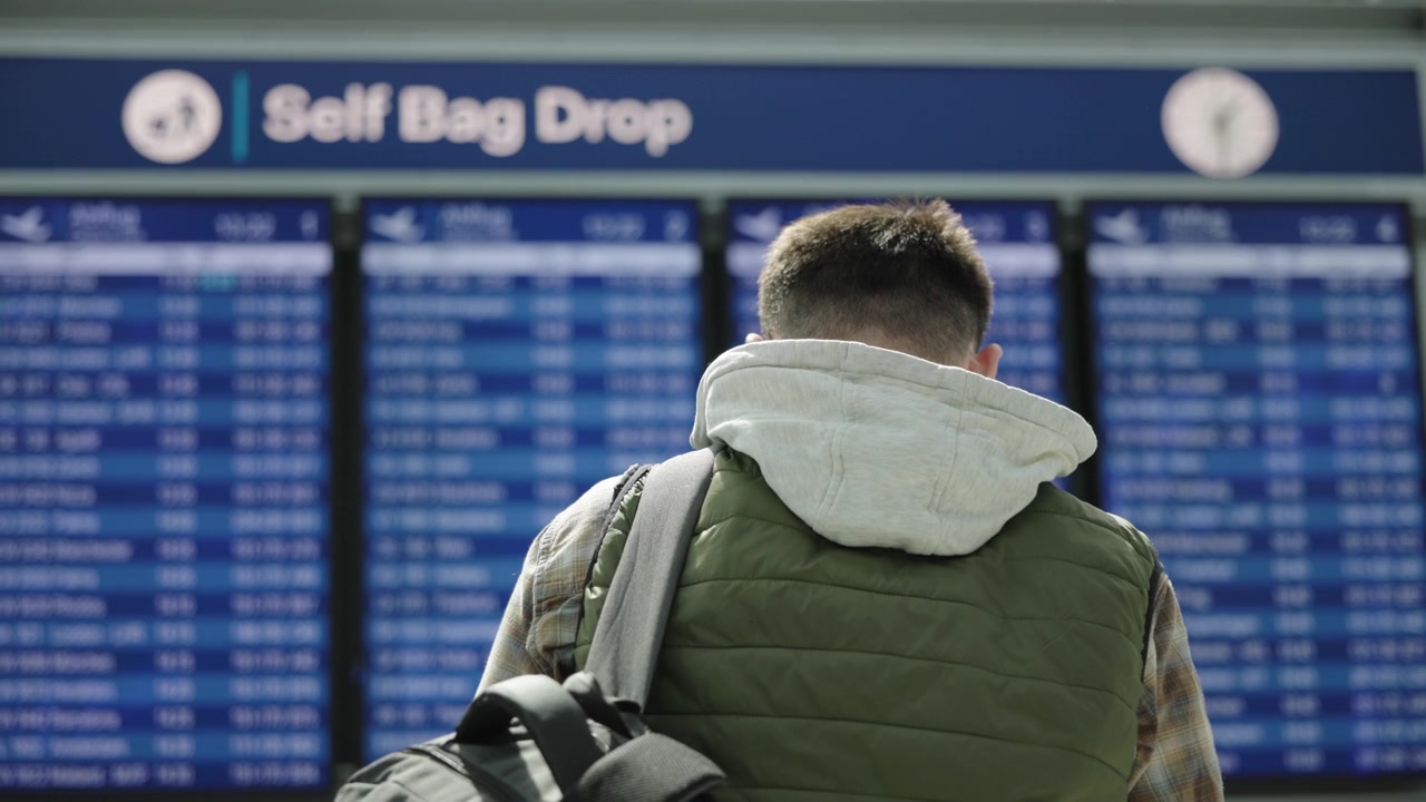 Giovane viaggiatore che guarda il tabellone delle partenze vicino all'area di consegna bagagli self-service in aeroporto