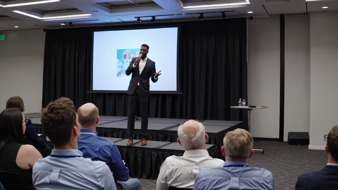 A speaker presents on stage in a conference room, captured from a rear audience angle