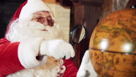 santa looking into a globe through the magnifying glass. portrait of old santa with a white beard wearing glasses looking for some place on a map in his residence