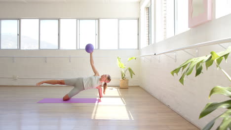 caucasian woman holding purple ball, doing yoga in sunny studio