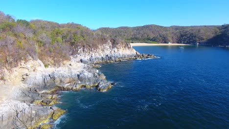 Forward-Strafe-Aerial-of-Rocky-Coast-to-Sailboat-Anchored-Near-Small-Beach-Cove