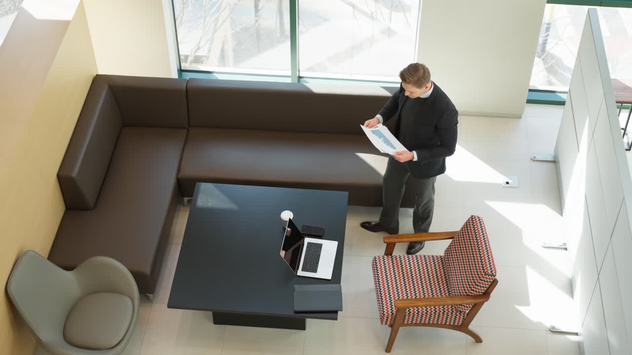 Caucasian business man stands in a modern office lounge holding reviewing paper documents - top down wide view