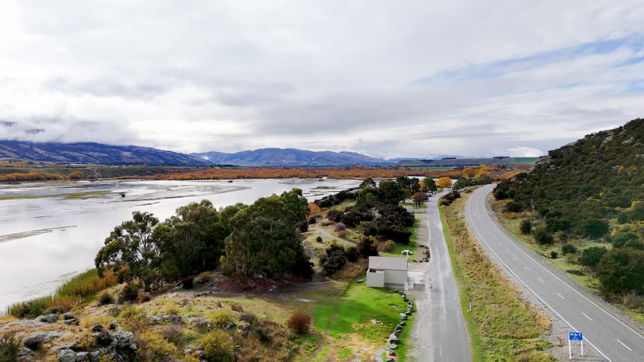 Drone footage glides above a winding lakeside road bordered by autumn trees, rocky hills, and reflective water under overcast skies, creating a tranquil rural scene