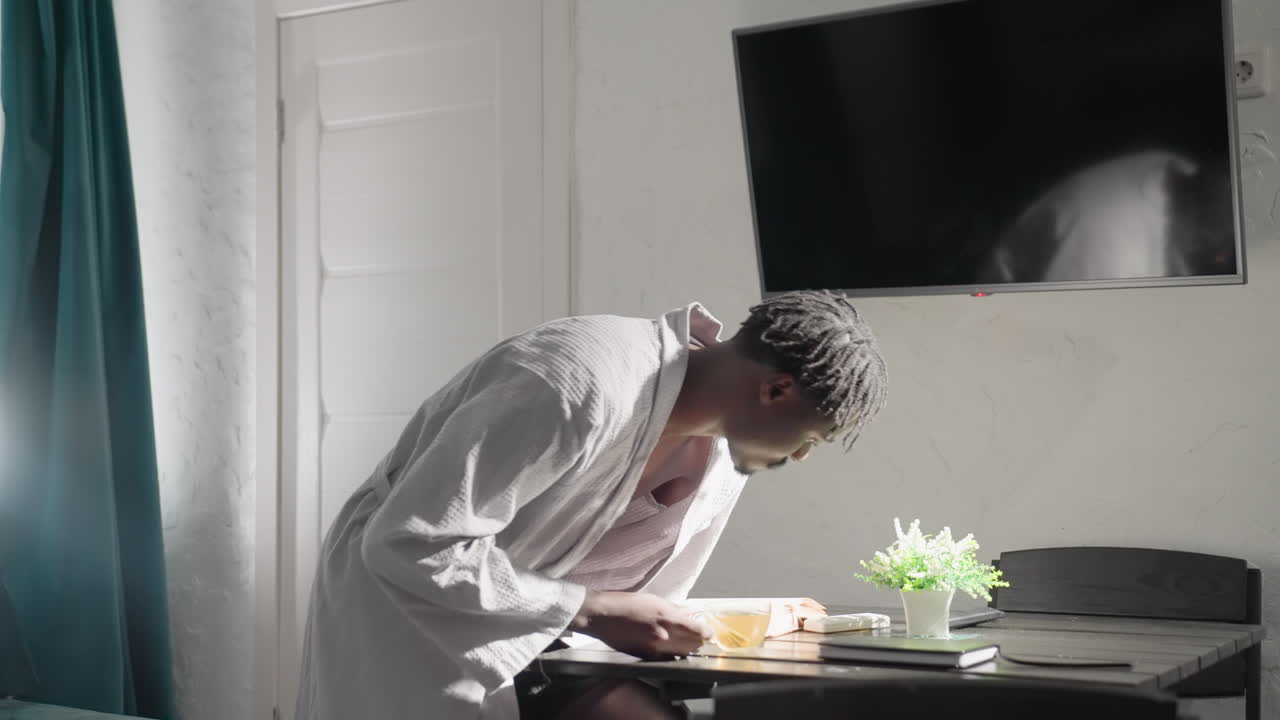 Man in white robe placing cup of tea on table in bright modern living space. Sunlight pours through window, illuminating peaceful, cozy atmosphere perfect for a quiet, relaxing moment