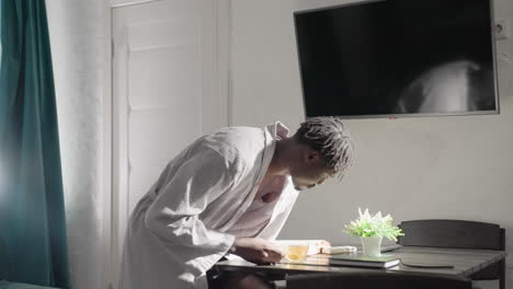 man in white robe placing cup of tea on table in bright modern living space. sunlight pours through window, illuminating peaceful, cozy atmosphere perfect for a quiet, relaxing moment
