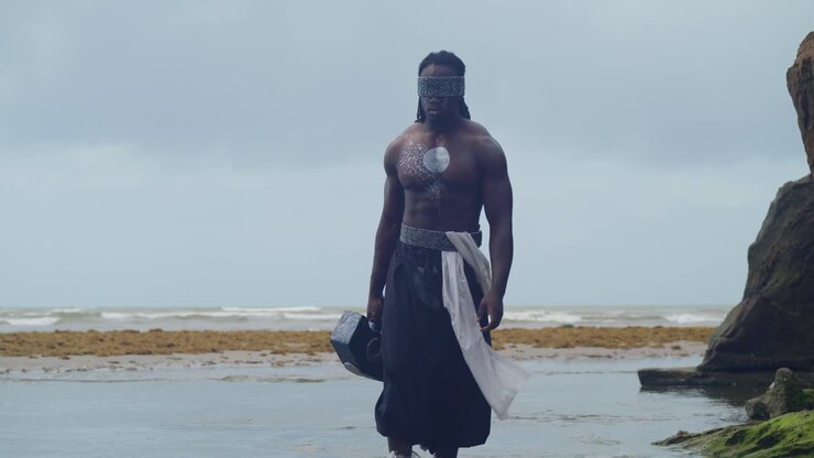 Su una spiaggia tropicale, un uomo muscoloso e dalla pelle scura interpreta Nekharis in cosplay in modo impeccabile.